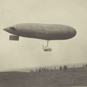 Ein historisches Foto eines großen Parseval Luftschiffs, das über einem Feld fliegt, mit Zuschauern, die unten versammelt sind.